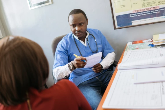 Foto dentro de um consultório hospitalar com um homem médico, negro, vestido de roupa azul e estetoscópio segura papéis na mão enquanto olha para uma pessoa a sua frente. A pessoa tem cabelo liso curto marrom e veste vermelho. Na mesa estão alguns papéis e na parede uma tela. 